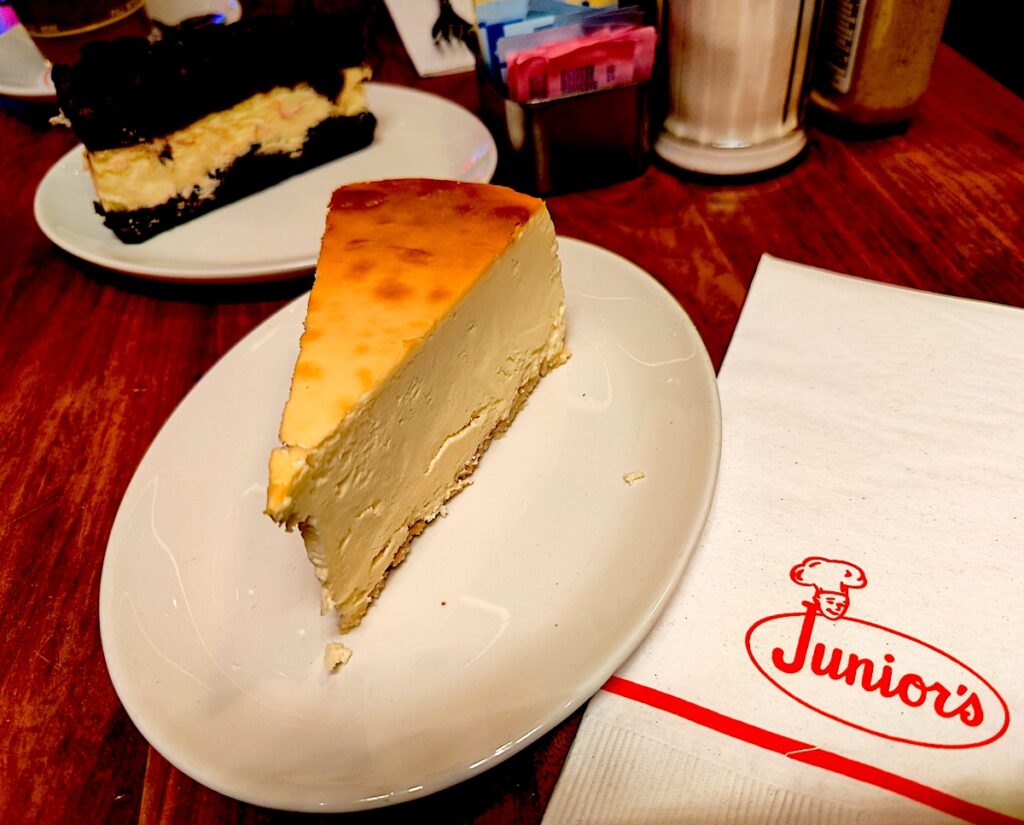 A piece of original cheesecake and brownie cheesecake from Juniors. There is a napkin to the right that says Junior's on it and the plates sit on a wooden table at one of the kid-friendly restaurants in NYC. 