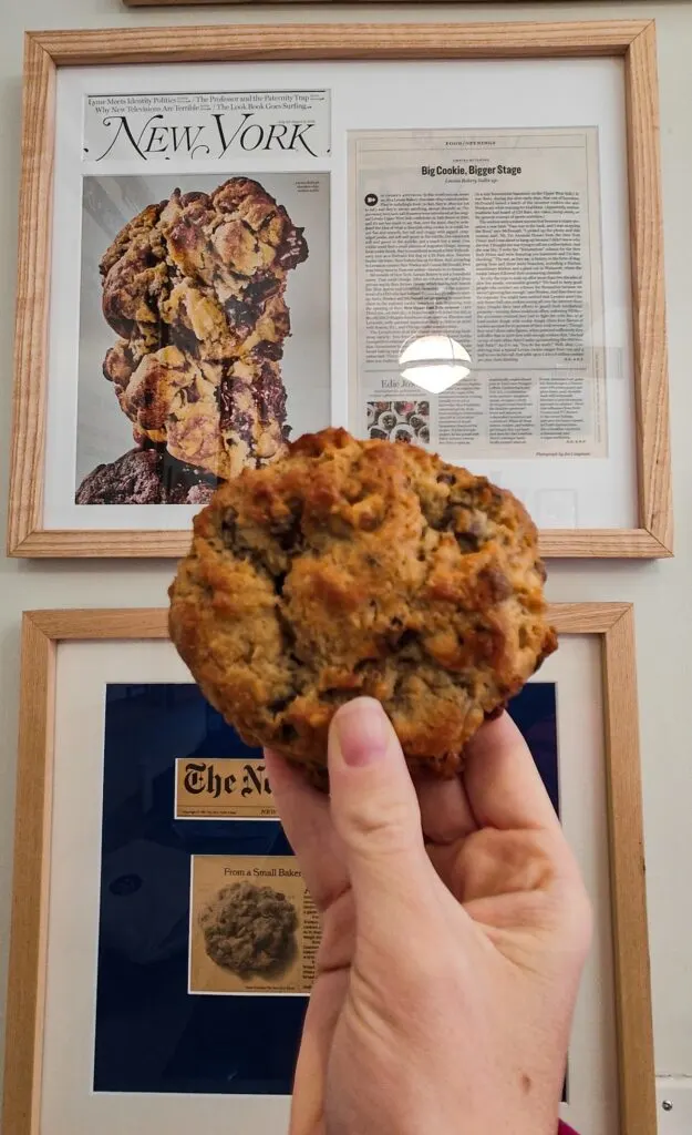 I am holding a chocolate chip cookie from Levain Bakery in front of framed articles about how amazing this cookie is. 