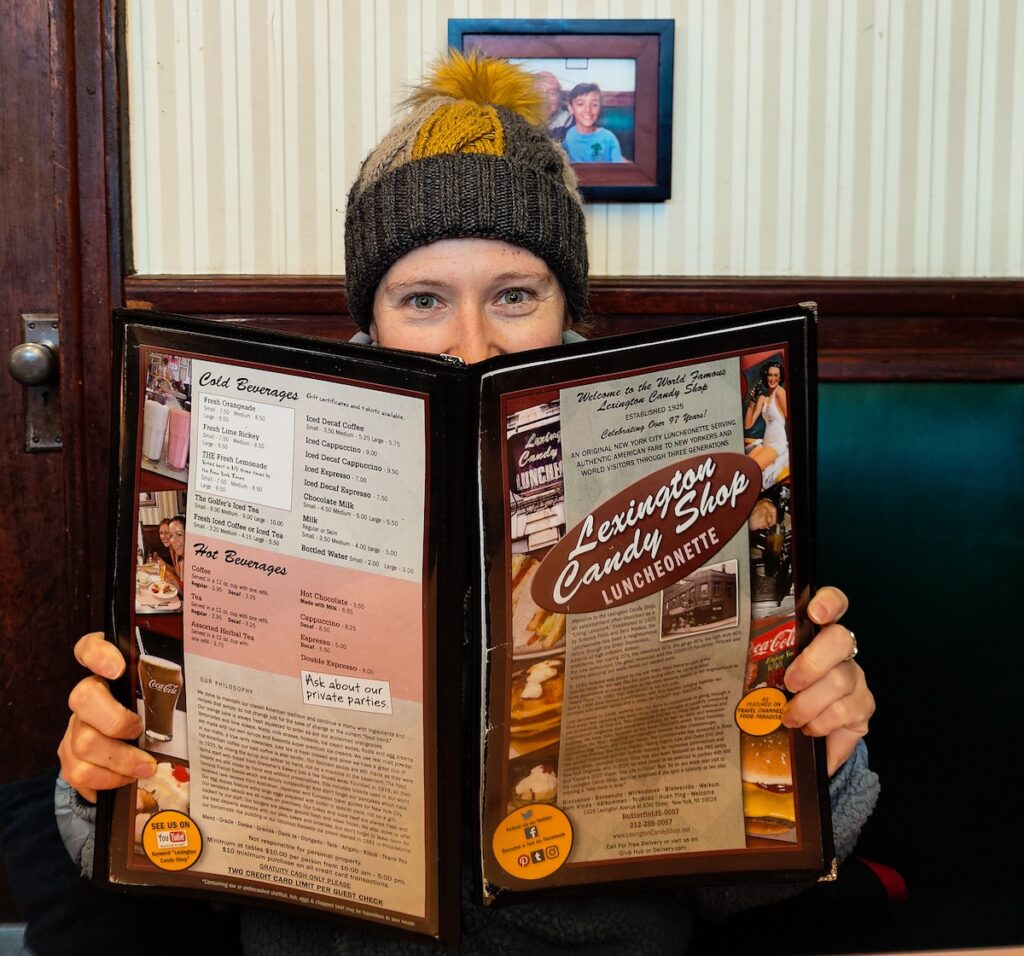 Me holding a menu for Lexington Candy Shop woth the vintage resaurant in the background. I am wearing a yellow and gray winter hat. 