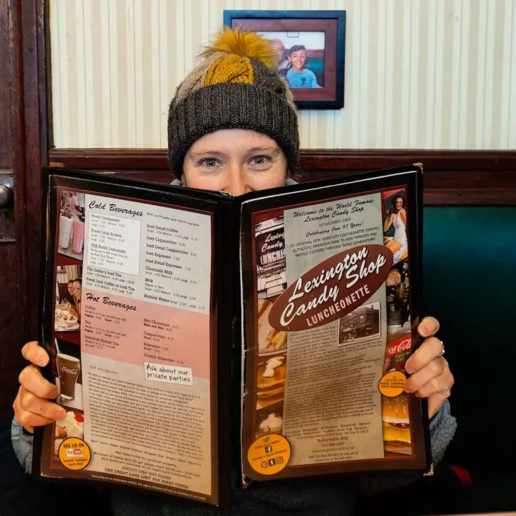 Me holding a menu for Lexington Candy Shop woth the vintage resaurant in the background. I am wearing a yellow and gray winter hat.