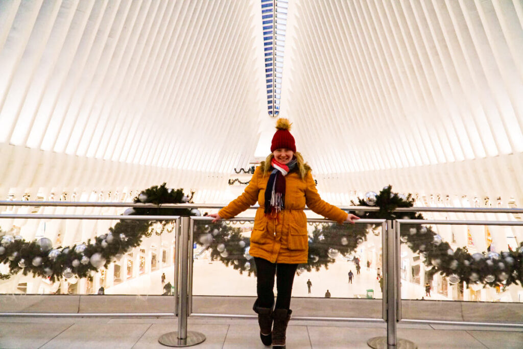 View of my standing pn the platform inside of the Oculus in New York City in February. I am wearing a mustard yellow coat, a plaid scarf, and a red hat and there is garland on the banister. 