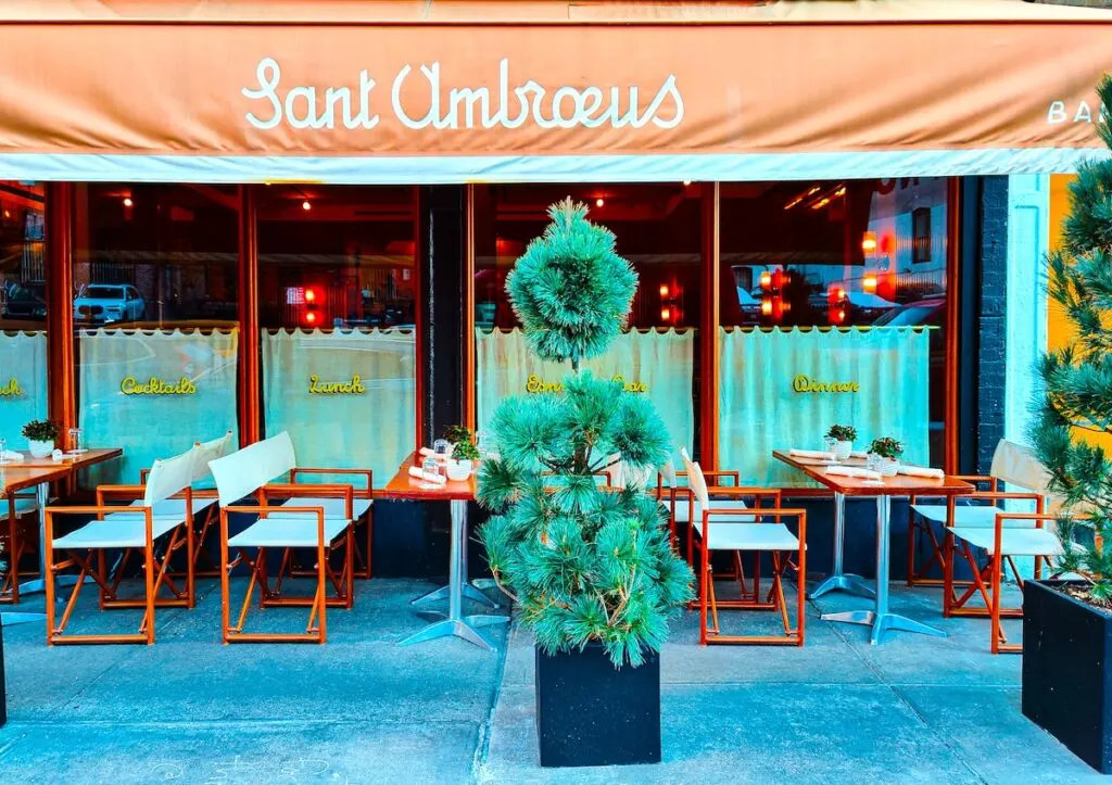 Exterior of Sant Ambroeus in NYC. There is a tan awning with the name of one of the best celebrity restaurants in NYC on it. You can see wooden tables and director-style chairs on the sidewalk with evergreen plants out front in black flower pots. 