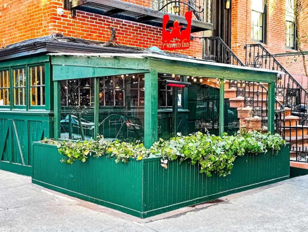 The exterior of the Waverly Inn sits on the ground floor of a historic, brick building. It is an enclosed green porch with plants that sit in window boxes. around the exterior of the restaurant. A red, 