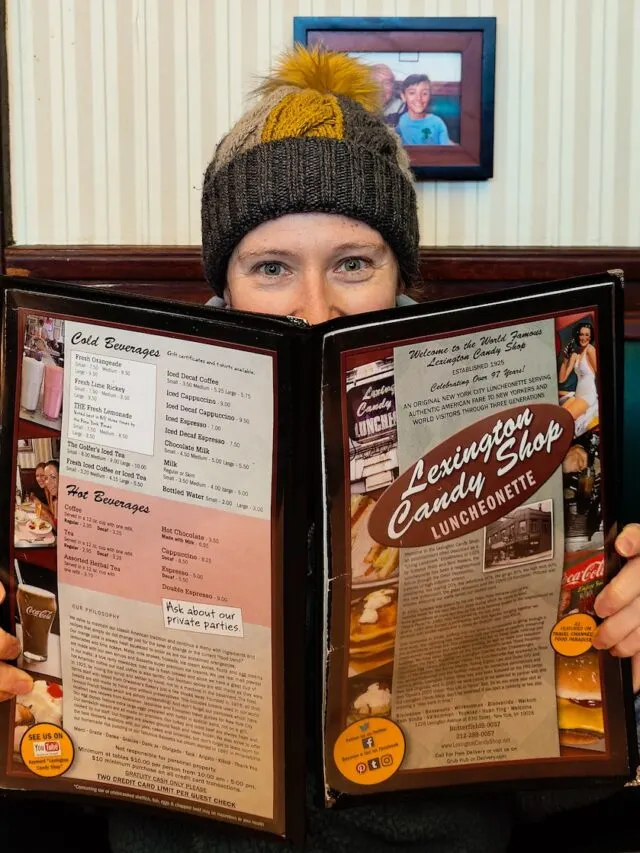 Me holding a menu for Lexington Candy Shop woth the vintage resaurant in the background. I am wearing a yellow and gray winter hat.