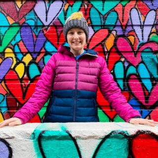 Me sitting in a pink and purple winter jacket with my hands on a white cement railing, I am looking at the camera and have a gray winter hat on with grafitti hearts in the background in NYC.