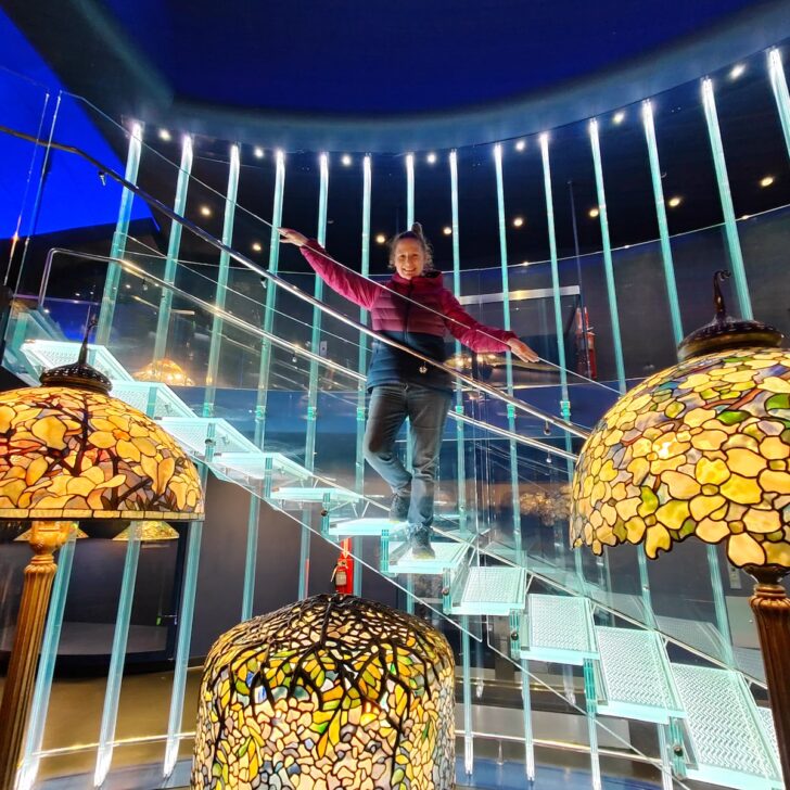View of me standing ona. glass stairway in front of three tiffany lamps on display in the New York Historical Society.