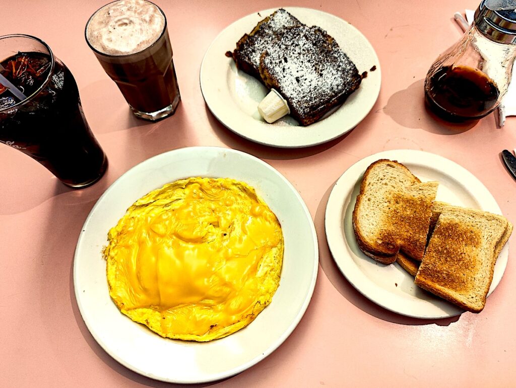 Aerial view of a cheese omelet, toast, french toast, and an egg cream from Lexington Candy Shop. The food all sits ona. pink counter. 