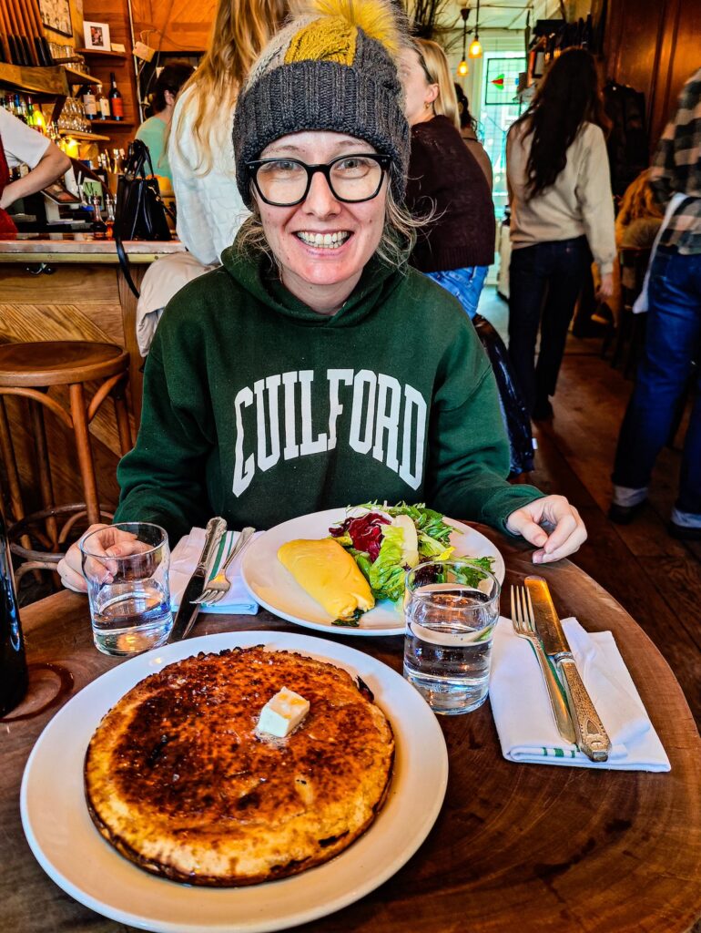 A view of Kelly sitting in a green sweatshirt and a gray winter hat with a plate with salad and eggs on it. There are also pancakes on the table in front of Kelly. 