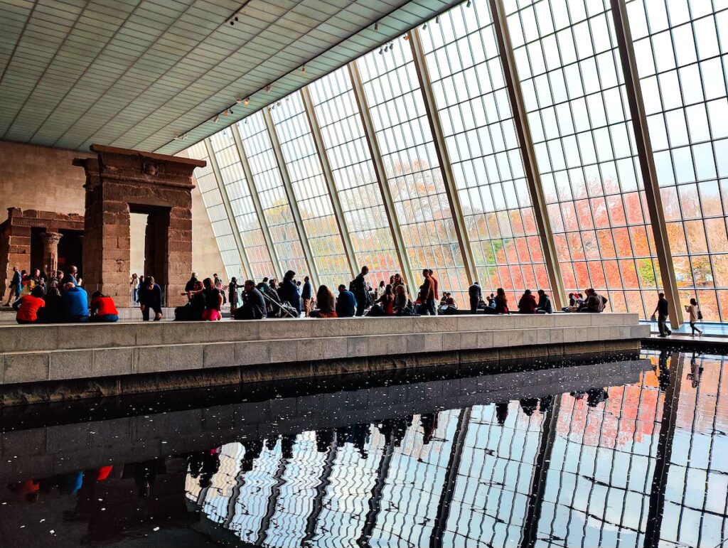 View of the Temple of Dendur at the Metropolitan Museum of Art. You can see a pool around the temple and slanted glass windows. 
