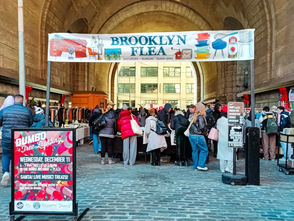 A view of people shopping beneath the Manhattan Bridge in Dumbo. You can see them standing beneath a huge sign that says, "Brooklyn Flea". 