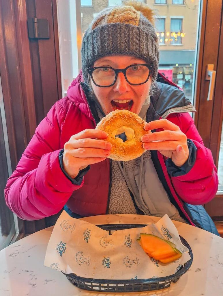 Me eating a Montreal-style sesame bagel with cream cheese. My moth is open and I am wearing a pink winter jacket while sitting at a table with a door in the background.  